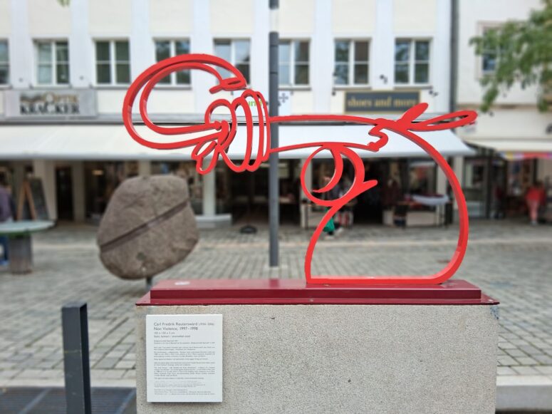 Eine rote Metallskulptur eines Revolvers, dessen Lauf verknotet ist, steht im Freien auf einem Sockel; CC0-Bilder. Dahinter steht ein großer Felsen auf einer Kopfsteinpflasterstraße, die im Hintergrund von Geschäften gesäumt ist.
