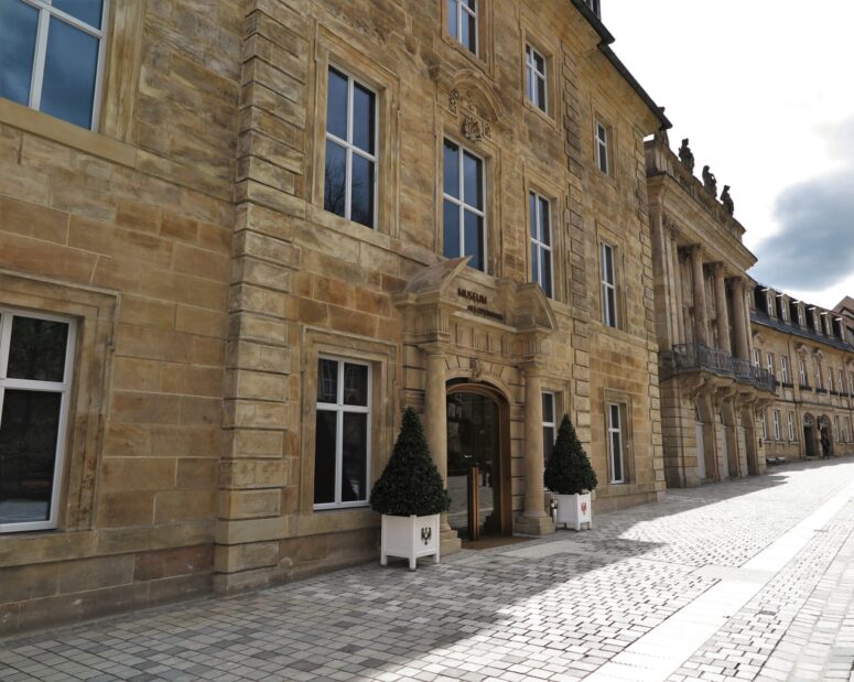 Das Bild, das als CC0-Bilder verfügbar ist, zeigt den Eingang zu einem Museum in einem historischen Steingebäude mit großen Fenstern, klassischen architektonischen Details und zwei Topfbäumen neben der Tür. Die Straße ist mit hellen Steinen gepflastert.