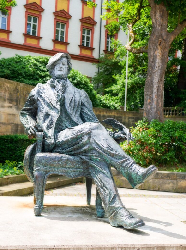 Die Bronzestatue eines sitzenden Mannes mit gekreuzten Beinen, der einen Hut trägt und einen Federkiel in der Hand hält, befindet sich im Freien auf einer gepflasterten Fläche in der Nähe von Bäumen und einem historischen Gebäude mit roten und weißen Wänden im Hintergrund.