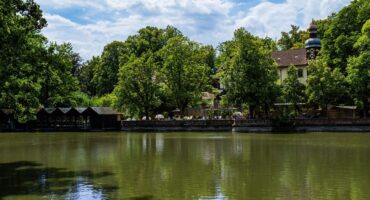 In einem ruhigen Teich spiegeln sich üppig grüne Bäume und ein traditionelles Gebäude mit einem Turm. Am Wasser sind Sitzgelegenheiten im Freien zu sehen, und über einem Teil des Teichs erstreckt sich eine Holzkonstruktion unter einem teilweise bewölkten Himmel, die in atemberaubenden CC0-Bildern festgehalten wurde.