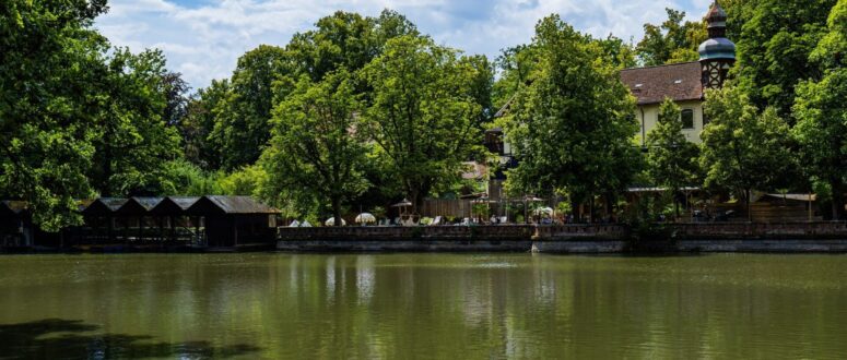In einem ruhigen Teich spiegeln sich üppig grüne Bäume und ein traditionelles Gebäude mit einem Turm. Am Wasser sind Sitzgelegenheiten im Freien zu sehen, und über einem Teil des Teichs erstreckt sich eine Holzkonstruktion unter einem teilweise bewölkten Himmel, die in atemberaubenden CC0-Bildern festgehalten wurde.