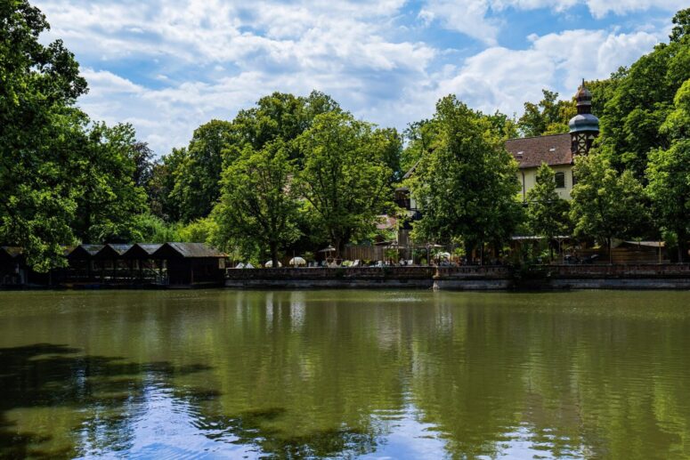 In einem ruhigen Teich spiegeln sich üppig grüne Bäume und ein traditionelles Gebäude mit einem Turm. Am Wasser sind Sitzgelegenheiten im Freien zu sehen, und über einem Teil des Teichs erstreckt sich eine Holzkonstruktion unter einem teilweise bewölkten Himmel, die in atemberaubenden CC0-Bildern festgehalten wurde.