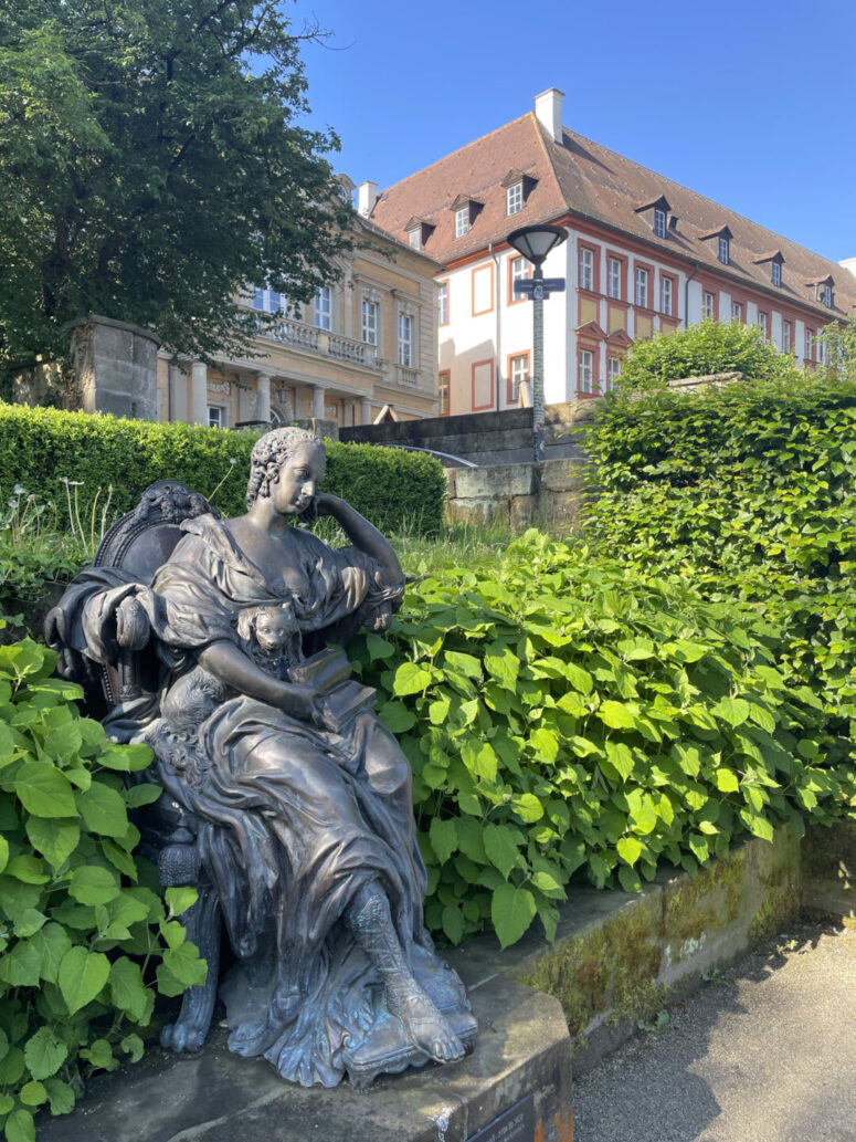 Eine Bronzestatue einer sitzenden Frau in einem wallenden Kleid ruht auf einem Steinsockel, umgeben von Grün, mit historischen Gebäuden und einer Straßenlaterne im Hintergrund unter einem klaren blauen Himmel - perfekt für CC0-Bilder-Sammlungen.