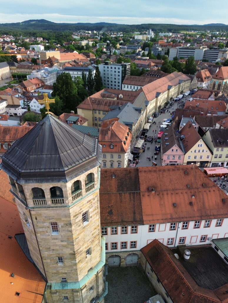 Luftaufnahme einer historischen europäischen Stadt mit roten Dächern, einem steinernen Kirchturm mit einem goldenen Kreuz und einem belebten Stadtplatz. Im Hintergrund erscheinen grüne Hügel und moderne Strukturen. Ideal für die Verwendung von CC0-Bildern.