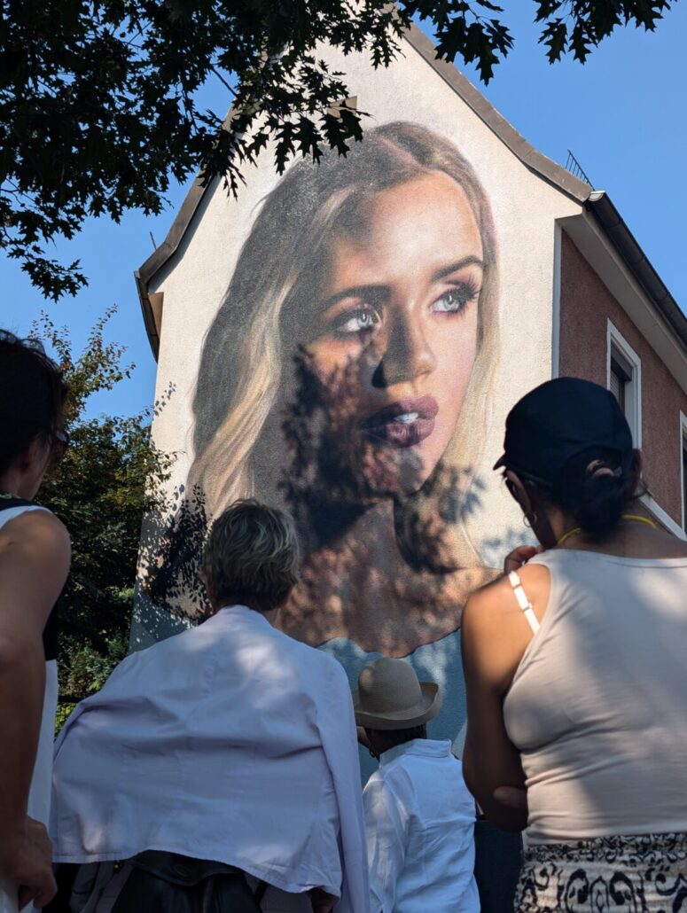 Eine Gruppe von Menschen steht vor einem Gebäude, auf dessen Seite ein großes, realistisches Wandgemälde eines Frauengesichts gemalt ist, während Sonnenlicht und Baumschatten auf die Szene fallen - perfekt für alle, die auffällige CC0-Bilder für kreative Projekte suchen.