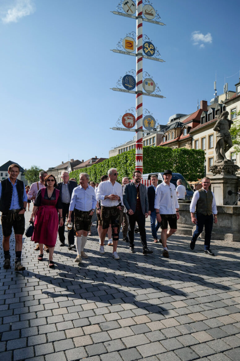 Eine Gruppe von Menschen, viele in traditioneller bayerischer Kleidung, spaziert an einem sonnigen Tag gemeinsam über einen Stadtplatz mit einem Maibaum und einer Statue im Hintergrund.