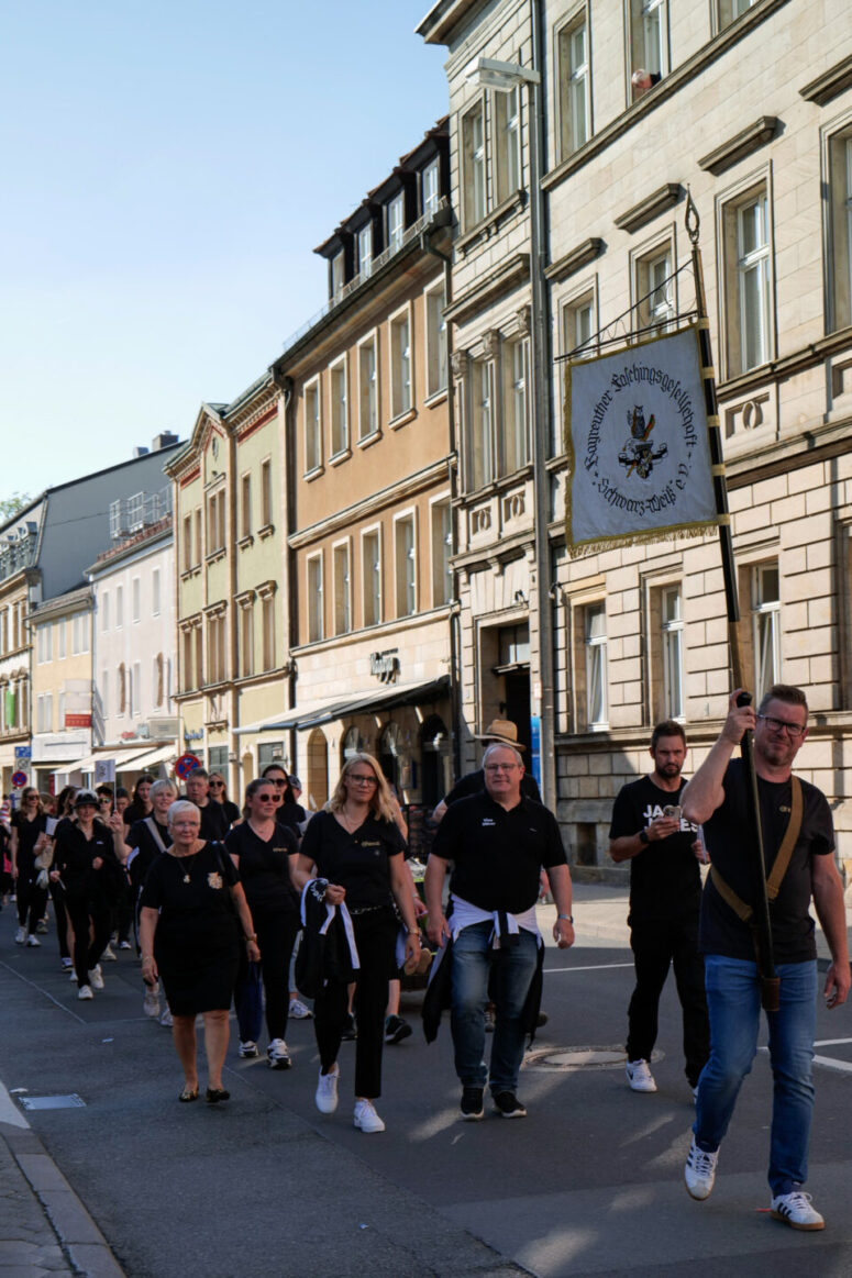 Eine Gruppe von meist schwarz gekleideten Menschen geht in einer Parade eine Straße entlang, angeführt von einem Mann, der ein Schild mit einem Emblem hält. Die Gebäude sind historisch und das Wetter ist sonnig.