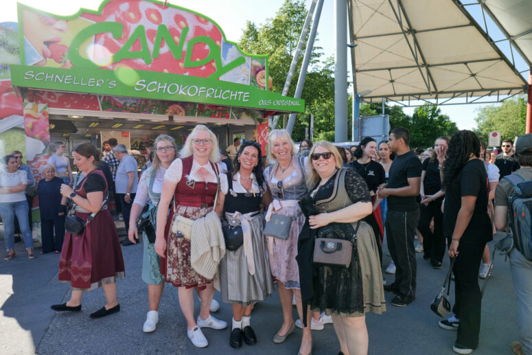 Eine Gruppe von Frauen in traditioneller bayerischer Kleidung lächelt und posiert gemeinsam vor einem bunten Süßigkeitenstand bei einer Veranstaltung im Freien, während im Hintergrund Menschen zusammenkommen und spazieren gehen.