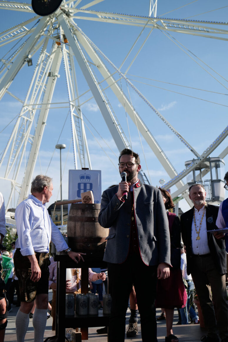 Ein Mann spricht in ein Mikrofon vor einem großen Riesenrad, neben einem Bierfass stehend und umgeben von Menschen in bayerischer Tracht. Die Szene suggeriert eine festliche Veranstaltung im Freien.