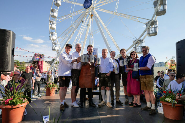 Eine Gruppe von Menschen in traditioneller bayerischer Kleidung steht auf einer Bühne mit Bierkrügen in der Hand, während im Hintergrund ein Riesenrad und eine jubelnde Menge auf einem Freiluftfestival zu sehen sind.