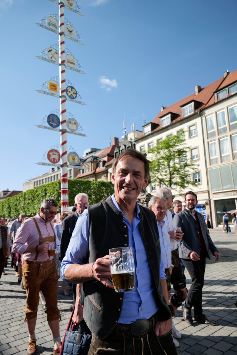 Ein lächelnder Mann in traditioneller bayerischer Kleidung hält einen Bierkrug bei einem Fest im Freien, mit einem Maibaum und einer Gruppe von Menschen in ähnlicher Kleidung hinter ihm, auf einem Stadtplatz an einem sonnigen Tag.