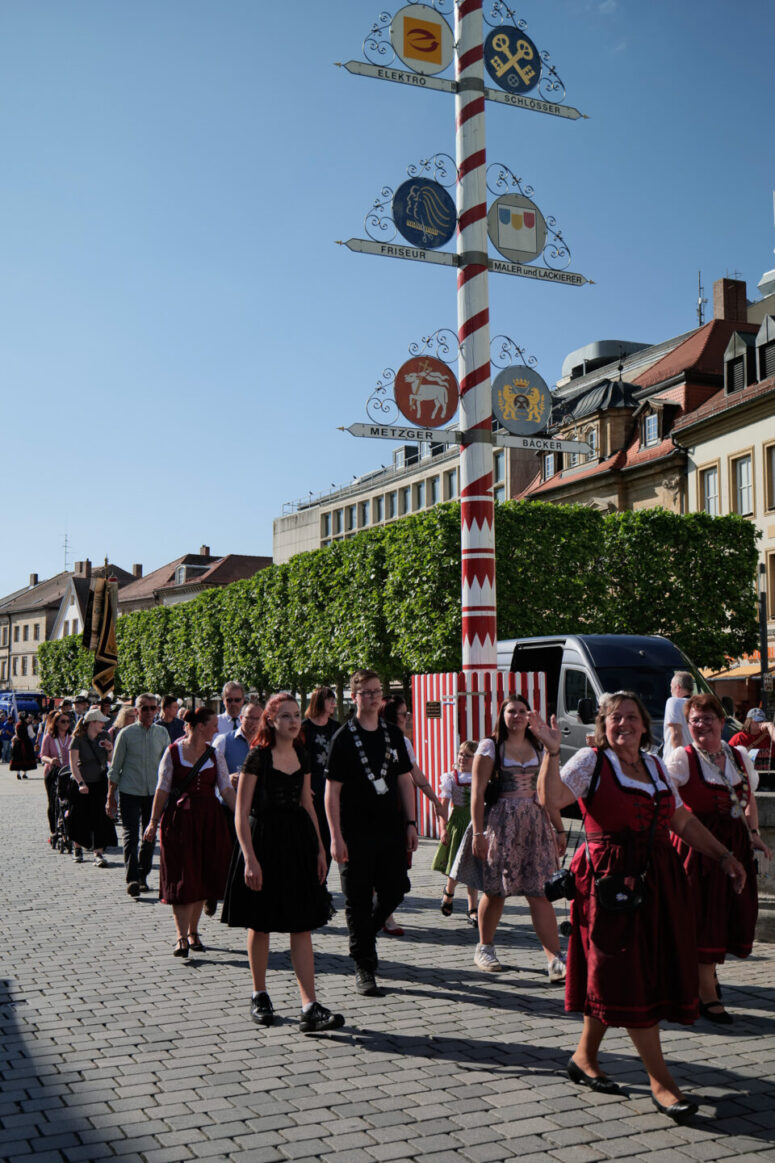 Eine Gruppe von Menschen in traditioneller Kleidung geht während eines Festes eine Kopfsteinpflasterstraße entlang, vorbei an einem hohen dekorativen Mast mit bunten Schildern. Gebäude und Bäume säumen den Hintergrund unter einem klaren blauen Himmel.