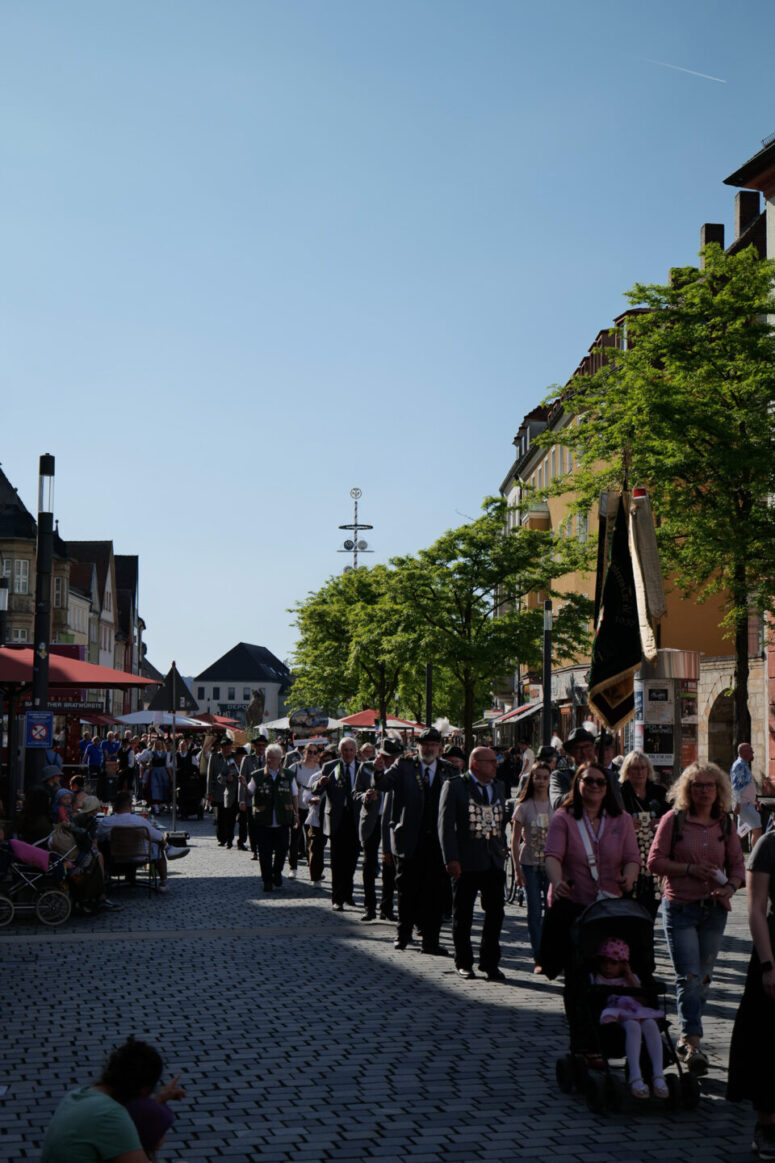 Eine Menschenmenge marschiert eine kopfsteingepflasterte Straße in einer sonnigen Stadt hinunter, einige tragen Transparente. Schaulustige schauen von den Seiten zu, und Bäume säumen die Straße unter einem klaren blauen Himmel.