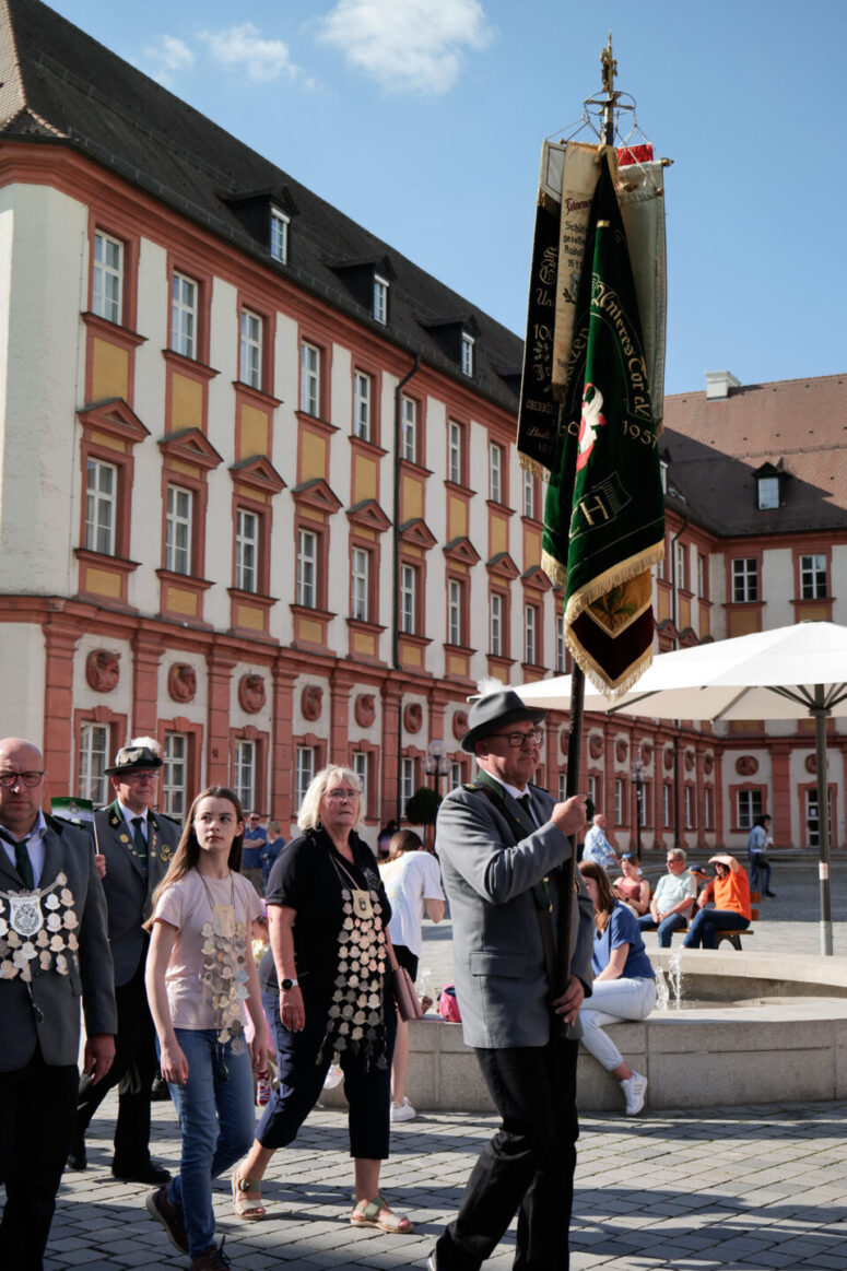 Eine Gruppe von Menschen, von denen einige Orden und Uniformen tragen, läuft in einer Parade. Ein Mann an der Spitze hält eine grüne Zeremonienfahne. Sie befinden sich auf einem historischen europäischen Platz mit einem großen, verschnörkelten Gebäude im Hintergrund.
