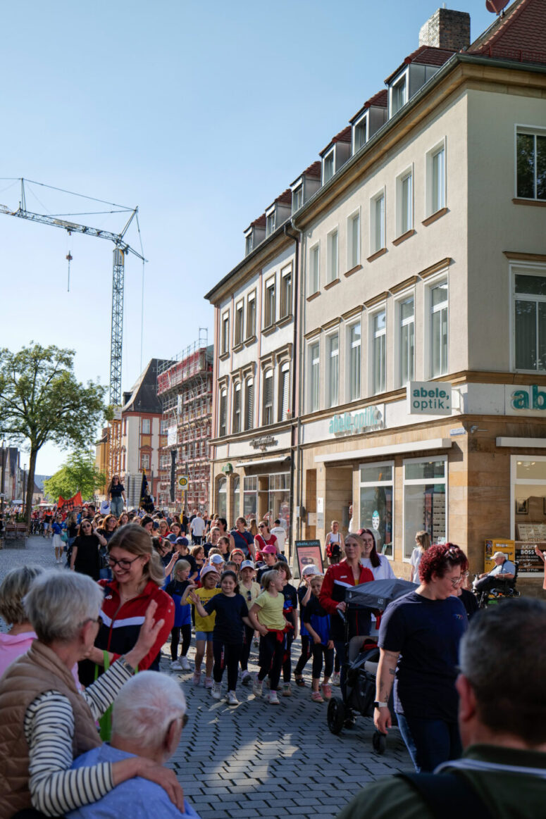 Eine lebhafte Straßenszene zeigt eine lange Gruppe von Kindern und Erwachsenen, die an einem sonnigen Tag gemeinsam an Geschäften und Schaulustigen vorbei eine Kopfsteinpflasterstraße in einer europäischen Stadt entlanggehen.