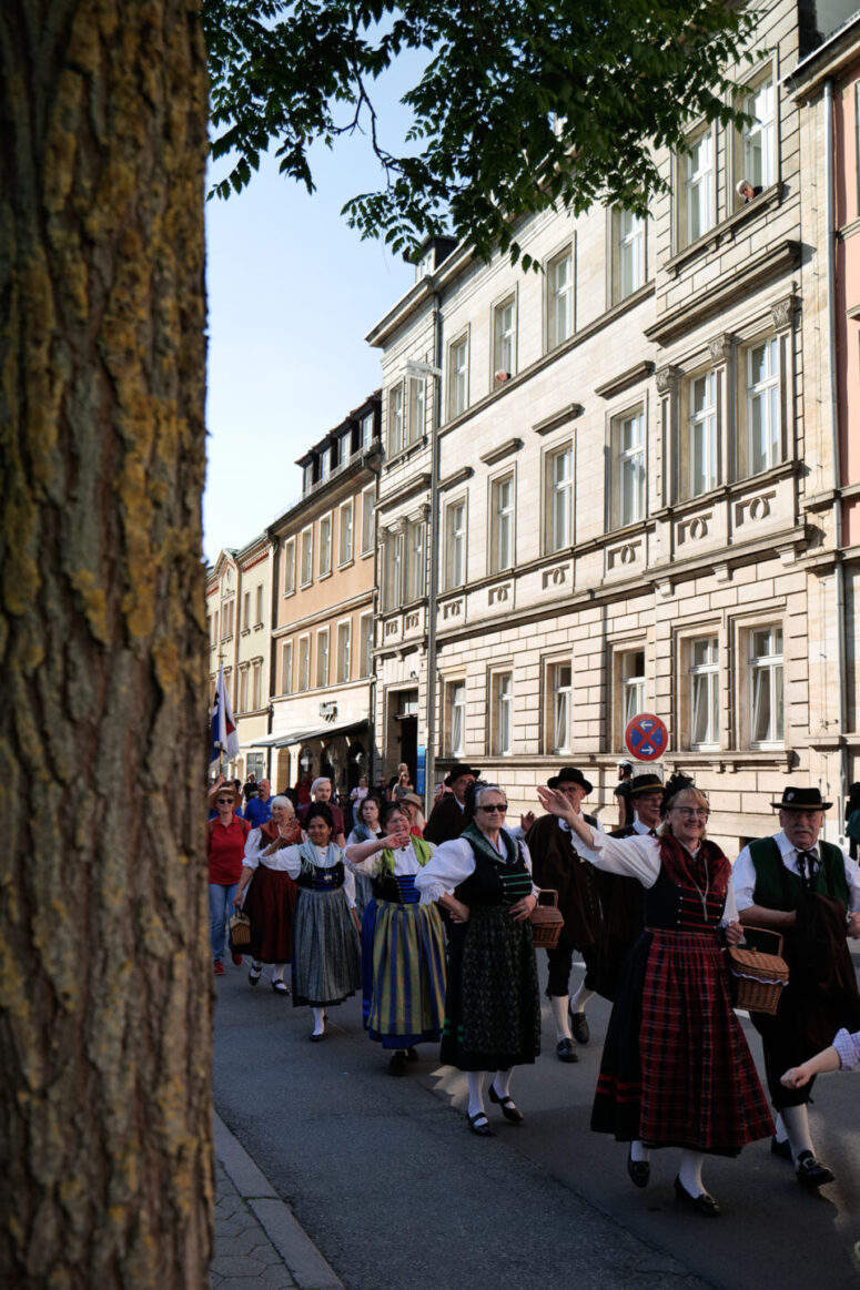 Menschen in traditioneller Kleidung gehen in einer festlichen Parade entlang einer von historischen Gebäuden gesäumten Stadtstraße. Ein großer Baumstamm ist im Vordergrund auf der linken Seite des Bildes zu sehen.