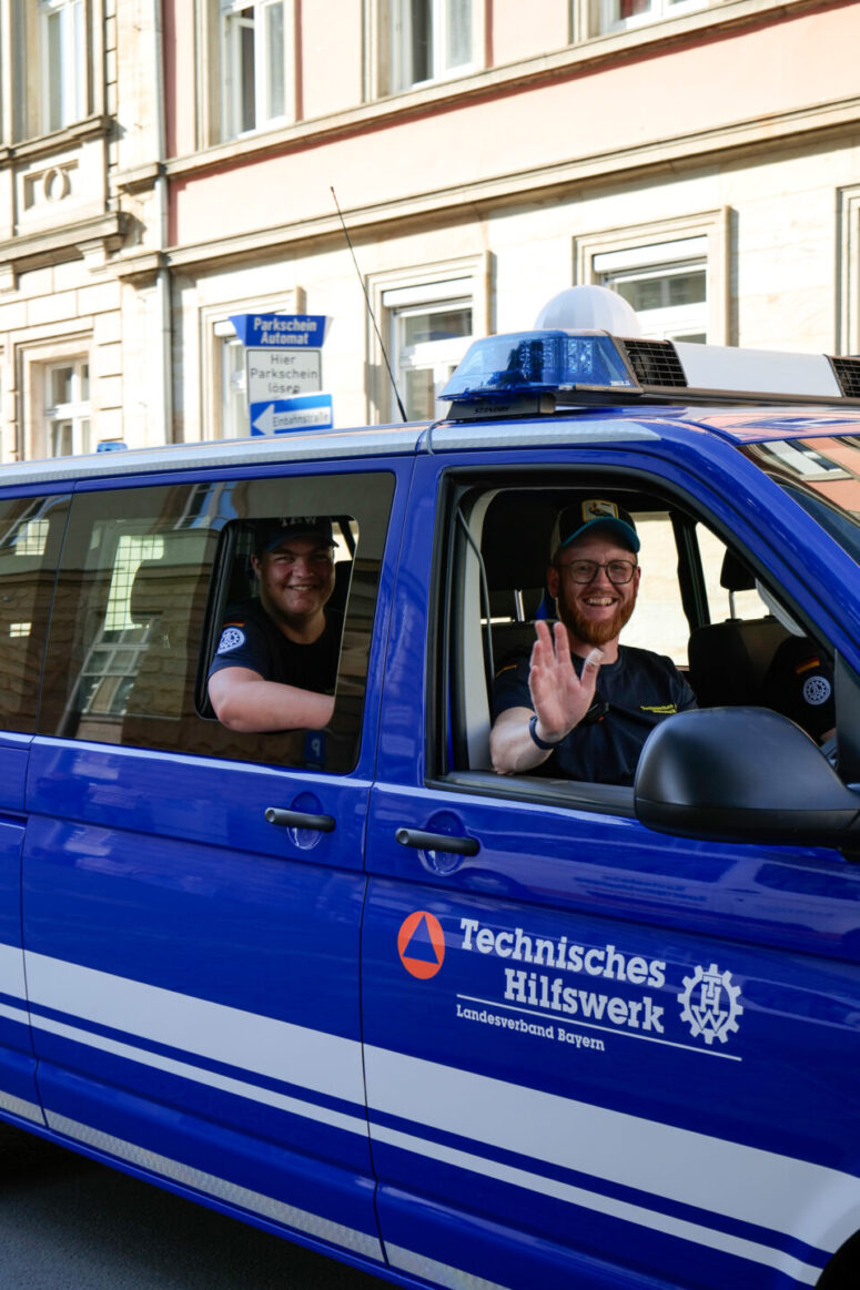 Zwei lächelnde Männer in Uniform sitzen in einem blauen Lieferwagen des Technischen Hilfswerks; einer winkt vom Beifahrersitz aus. Das Fahrzeug ist auf einer Straße vor Gebäuden geparkt, im Hintergrund ist ein Straßenschild zu sehen.