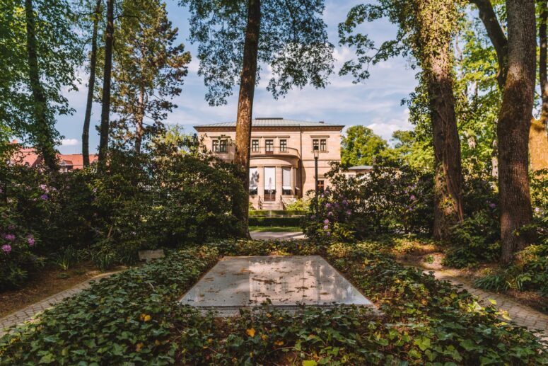 Im Vordergrund steht ein sonnenbeschienenes, von Efeu und Grün umgebenes Steinmonument, während im Hintergrund ein historisches Gebäude mit hohen Bögen und Säulen durch die Bäume hindurch sichtbar ist.
