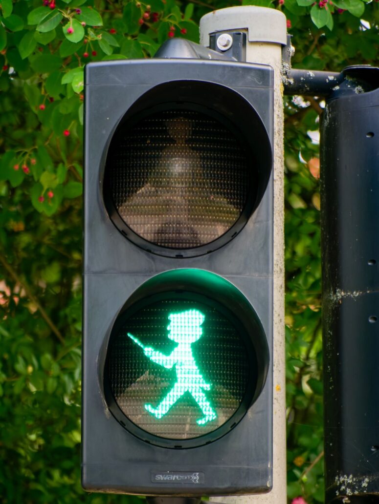 Eine Fußgängerampel mit einer grün leuchtenden Fußgängerfigur, die signalisiert, dass das Überqueren sicher ist. Im Hintergrund ist eine Grünfläche zu sehen.