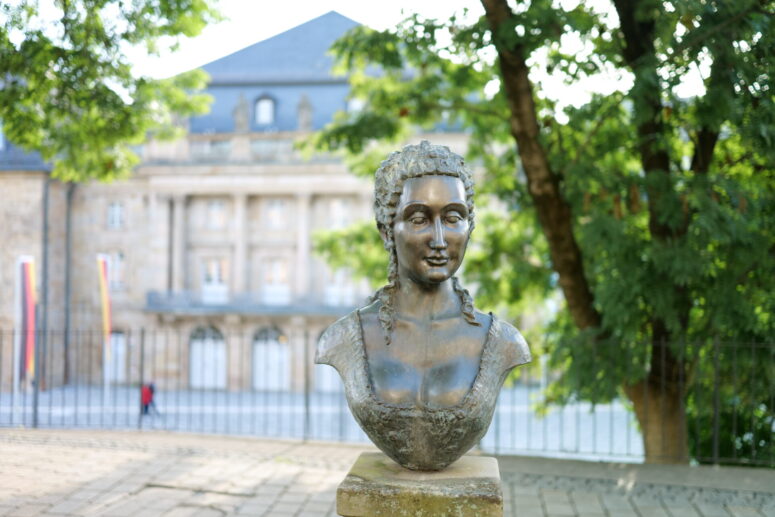 Eine Bronzebüste einer Frau mit geflochtenem Haar steht im Freien auf einem Sockel, eingerahmt von grünen Bäumen und einem historischen Steingebäude. Dieses Bild, das Teil der CC0-Bilder-Sammlung ist, kann für jeden Zweck frei verwendet werden.