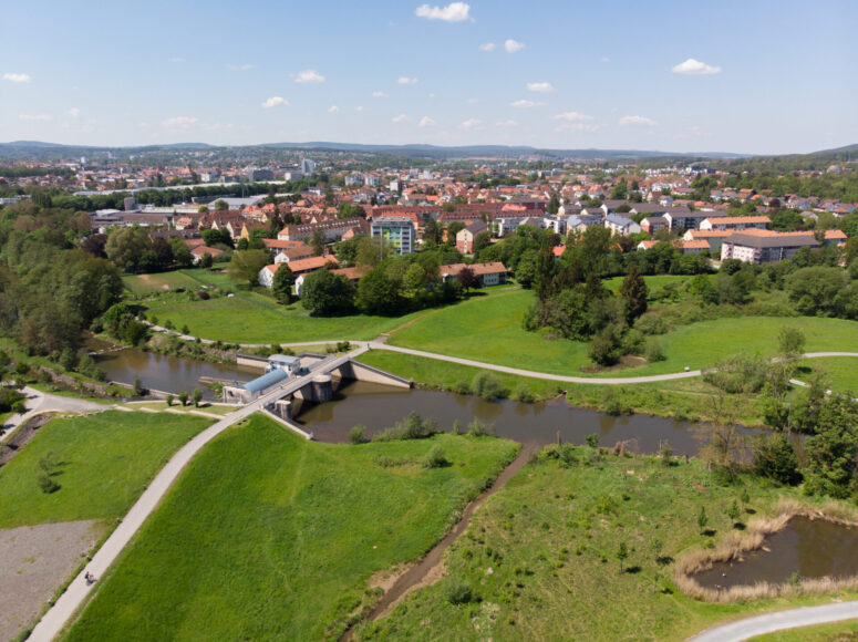 Luftaufnahme einer kleinen Stadt mit roten Dächern, grünen Feldern und Bäumen. Ein Fluss fließt durch die Szene mit einer Fußgängerbrücke und Wegen, die ihn unter einem klaren, sonnigen Himmel überqueren - eine einladende CC0-Bilder-Landschaft.