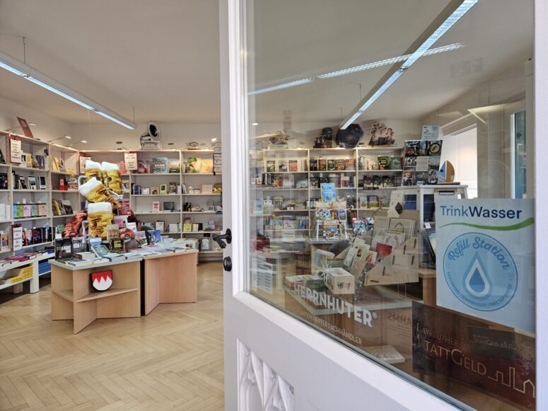 Das Innere einer Buchhandlung mit Holzböden, Regalen voller Bücher und einem Ausstellungsstand in der Mitte. An der Tür hängt ein Schild mit der Aufschrift „TrinkWasser-Auffüllstation“. Der Raum ist dank großer Fenster hell beleuchtet.