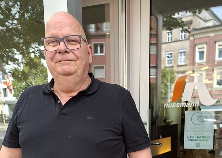 Ein glatzköpfiger Mann mit Brille, der ein schwarzes Poloshirt trägt, steht vor einem Gebäude mit Glastüren, auf denen das Nussmann-Logo und mehrere Schilder in deutscher Sprache zu sehen sind. Im Hintergrund sind Wohnhäuser und Bäume zu erkennen.