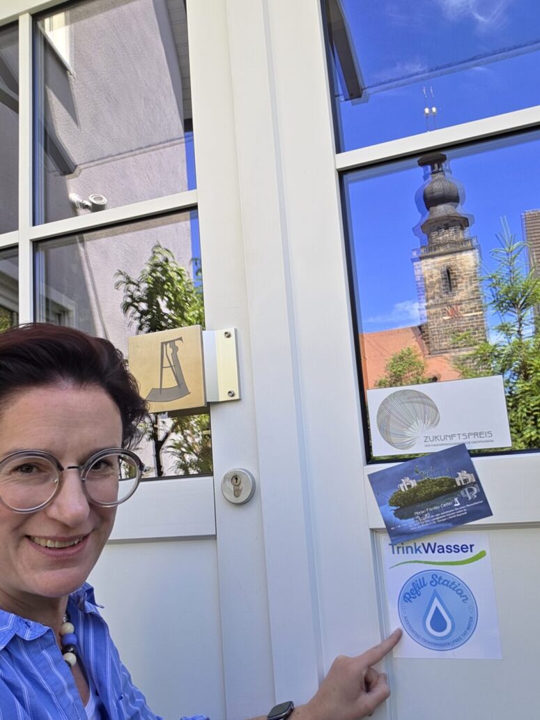 Eine Frau mit Brille und kurzen Haaren zeigt auf einen „Refill Station“-Aufkleber an einer weißen Tür. An der Tür befinden sich weitere Aufkleber, und in der Glasscheibe spiegeln sich ein Kirchturm, Bäume und Gebäude.
