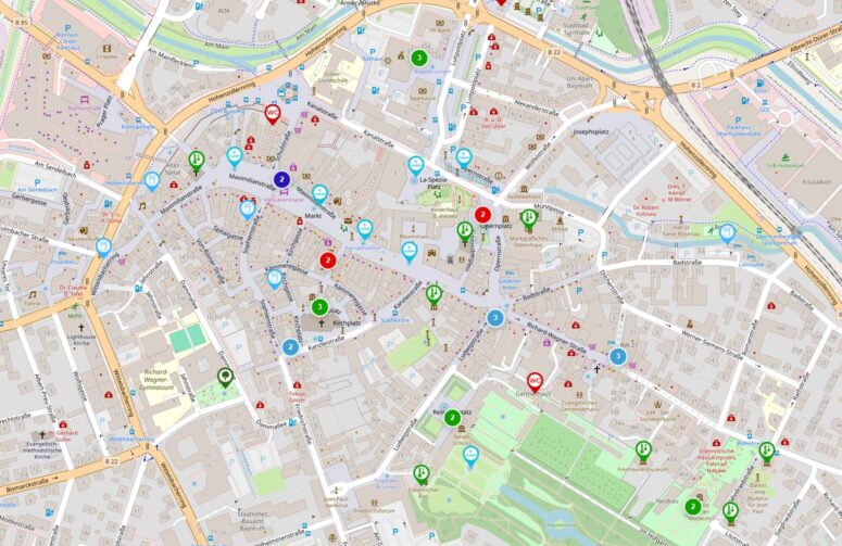 Ein detaillierter Stadtplan zeigt Straßen, Parks und Gebäude, die mit verschiedenfarbigen Symbolen für Einrichtungen wie Restaurants, Sehenswürdigkeiten, öffentliche Verkehrsmittel, Apotheken, Banken, Toiletten und Informationsstellen gekennzeichnet sind.