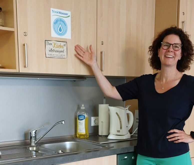 Eine lächelnde Frau mit lockigem Haar und Brille deutet auf ein Schild über einem Spülbecken, auf dem „Trinkwasser“ steht. Spülmittel, ein Wasserkocher und andere Küchenutensilien stehen auf der Arbeitsplatte neben ihr.