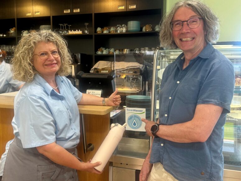 Zwei Menschen sitzen lächelnd in einem Café. Der eine hält eine wiederverwendbare Wasserflasche in der Nähe der Theke in der Hand, während der andere über einem Stapel Tassen ein Schild mit der Aufschrift „Trinkwasser“ hochhält. Im Hintergrund sind Regale mit Geschirr zu sehen.