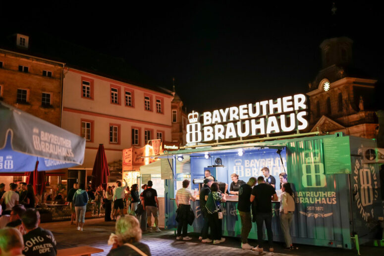 Während des Bürgerfestes 2026 versammeln sich die Menschen nachts an einem hell erleuchteten Bierstand des Bayreuther Brauhauses auf einem Platz im Freien, umgeben von historischen Gebäuden und beleuchteten Schildern.