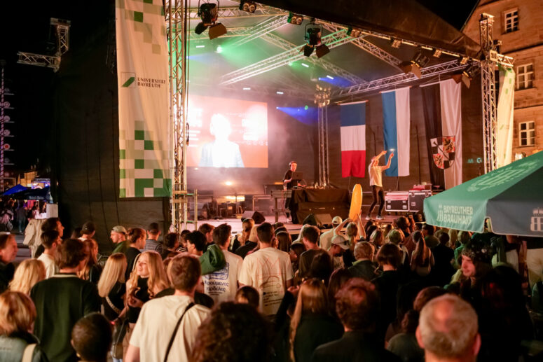 Eine große Menschenmenge verfolgt nachts während des Bürgerfestes 2026 den Auftritt eines DJs auf einer Außenbühne, im Hintergrund sind bunte Lichter, Fahnen und eine Leinwand zu sehen. Die Veranstaltung wirkt festlich und lebhaft.
