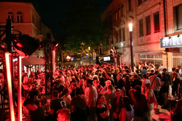 Eine große Menschenmenge versammelt sich nachts im Freien auf einer Straße der Stadt zum Bürgerfest 2026, das von rotem Licht beleuchtet wird; im Hintergrund sind Gebäude und ein SPIEL-Schild zu sehen.