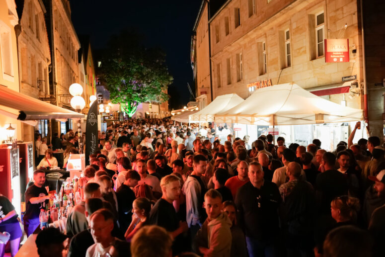 Während des Bürgerfestes 2026 versammelt sich nachts eine große Menschenmenge auf einem hell erleuchteten Straßenmarkt, umgeben von Ständen, Zelten und historisch anmutenden Gebäuden. Einige Leute bestellen an einem Essens- oder Getränkestand.