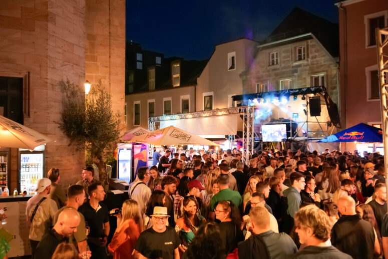 Eine große Menschenmenge genießt das Bürgerfest 2026, ein nächtliches Open-Air-Fest auf einem Stadtplatz, umgeben von Gebäuden und Imbissständen, mit Beleuchtung und einer Bühne im Hintergrund.