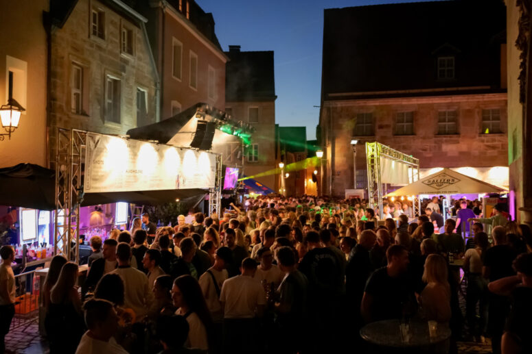 Bei einem Nachtfest im Freien im Rahmen des Bürgerfestes 2026 versammelt sich eine große Menschenmenge auf einem historischen Stadtplatz, der von alten Gebäuden umgeben und von bunten Lichtern beleuchtet ist und auf dem sich Essensstände und gesellige Menschen tummeln.