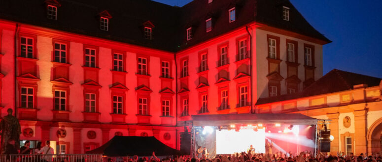 Eine große Menschenmenge genießt während des Bürgerfestes 2026 ein nächtliches Open-Air-Konzert vor einem historischen Gebäude, das in rotes Licht getaucht ist. Eine Bühne mit Künstlern ist zu sehen, und die Menschen tanzen und feiern.