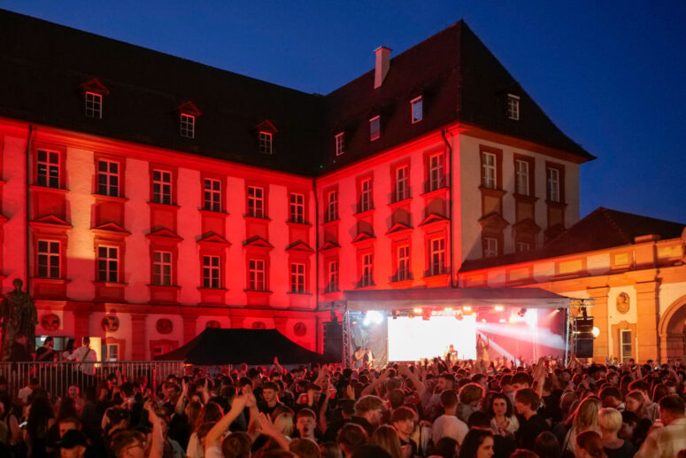Eine große Menschenmenge genießt während des Bürgerfestes 2026 ein nächtliches Open-Air-Konzert vor einem historischen Gebäude, das in rotes Licht getaucht ist. Eine Bühne mit Künstlern ist zu sehen, und die Menschen tanzen und feiern.