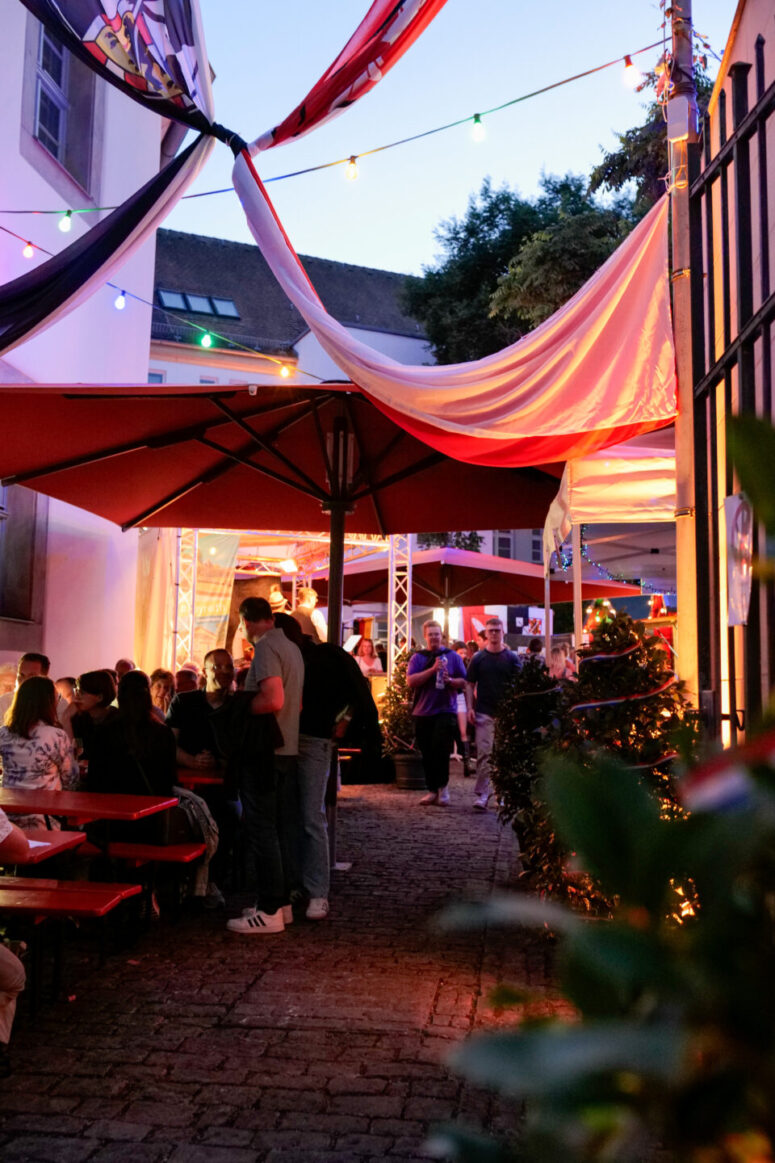 Eine lebhafte Szene im Freien in der Abenddämmerung zeigt Menschen, die sich während des Bürgerfestes 2026 an Tischen unter Markisen und Lichterketten unterhalten, wobei rote Sonnenschirme und festliche Dekorationen die gemütliche Atmosphäre im Innenhof unterstreichen.