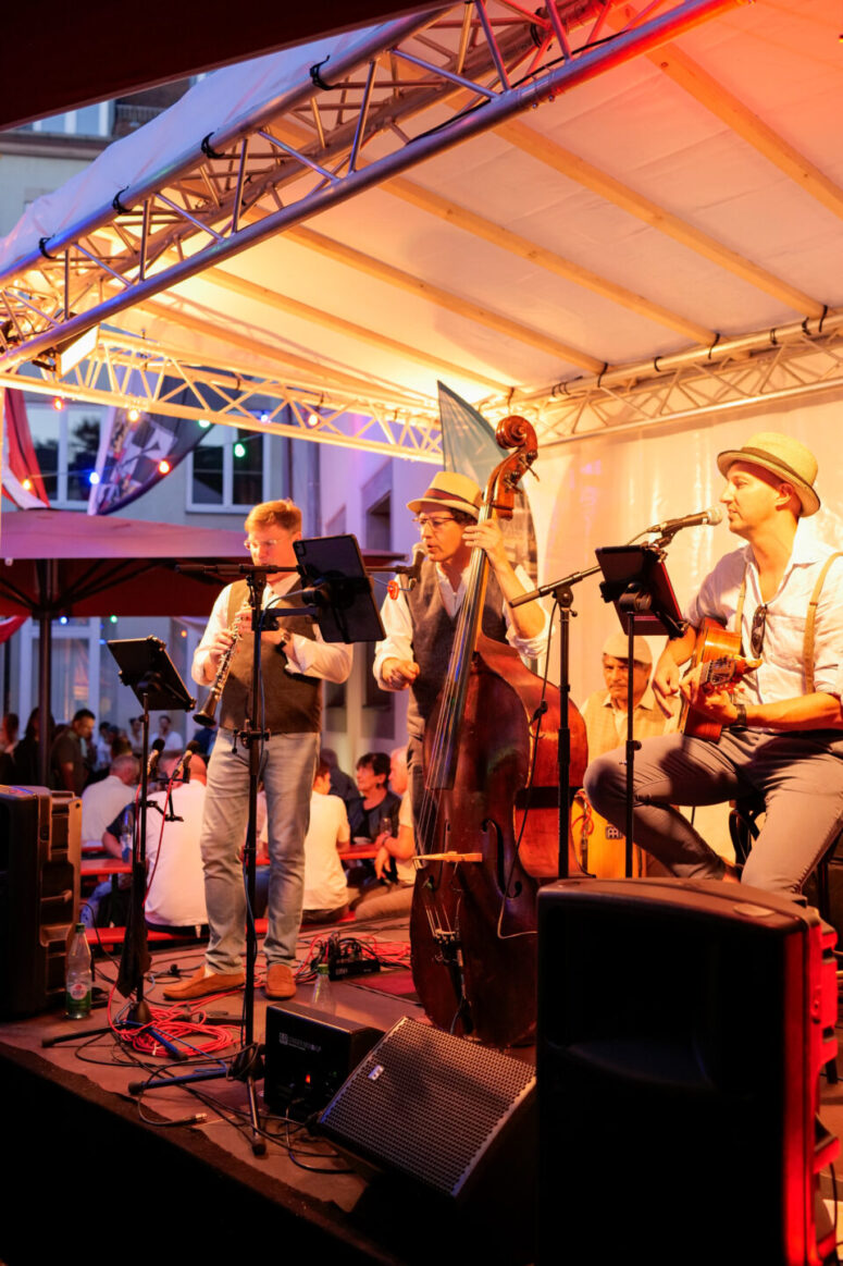 Drei Musiker treten während des Bürgerfestes 2026 nachts auf einer Freiluftbühne auf und spielen Klarinette, Kontrabass und Gitarre. Sie tragen Hüte und Freizeitkleidung. Warmes Licht beleuchtet sie, während im Hintergrund Menschen sitzen und zuschauen.