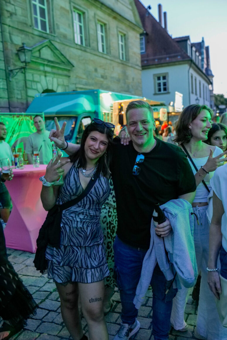 Zwei Menschen lächeln und zeigen das Friedenszeichen, während sie gemeinsam auf dem Bürgerfest 2026 posieren. Im Hintergrund sind weitere Menschen und bunte Foodtrucks zu sehen, umgeben von historischen Gebäuden. Das Abendlicht sorgt für eine festliche Atmosphäre.