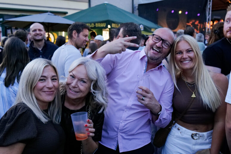 Vier Erwachsene lächeln und posieren auf dem Bürgerfest 2026. Ein Mann mit Brille und rosa Hemd zeigt das Peace-Zeichen, während er ein Getränk in der Hand hält. Drei Frauen stehen dicht beieinander und lächeln alle. Menschenmengen und Zelte prägen den Hintergrund dieser lebhaften Freiluftveranstaltung.