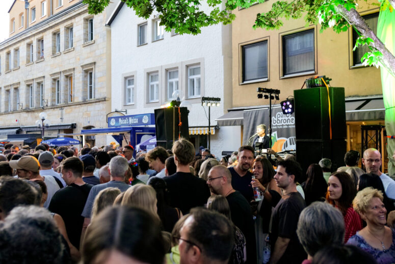 Bei dem Bürgerfest 2026 versammelt sich eine große Menschenmenge bei einem Straßenfest im Freien vor Geschäften und Gebäuden und genießt im Abendlicht die Live-Musik, die auf einer kleinen Bühne unter laubreichen Bäumen gespielt wird.
