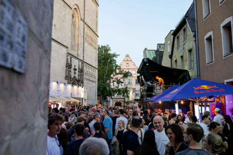 Beim Bürgerfest 2026, einem Straßenfest unter freiem Himmel inmitten historischer Gebäude mit Imbissständen und einem Red-Bull-Zelt, versammelt sich eine große Menschenmenge. Auf der Bühne legt ein DJ auf, während die Besucher die lebhafte Atmosphäre genießen.