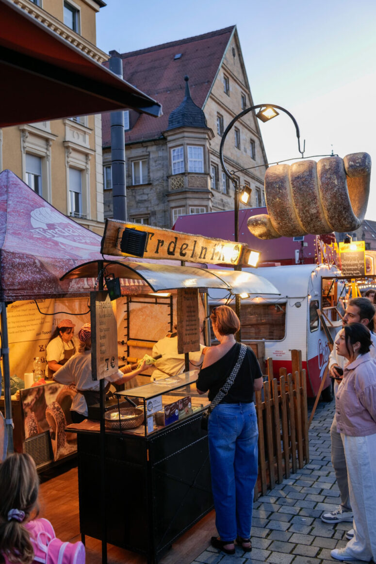 Auf dem Bürgerfest 2026 stehen Menschen an einem Marktstand im Freien Schlange, an dem Lebensmittel verkauft werden; über dem Stand hängt ein großes Zierbrot. Im Inneren bereitet der Verkäufer Gebäck zu, und im Hintergrund sind bei Sonnenuntergang historische Gebäude zu sehen.