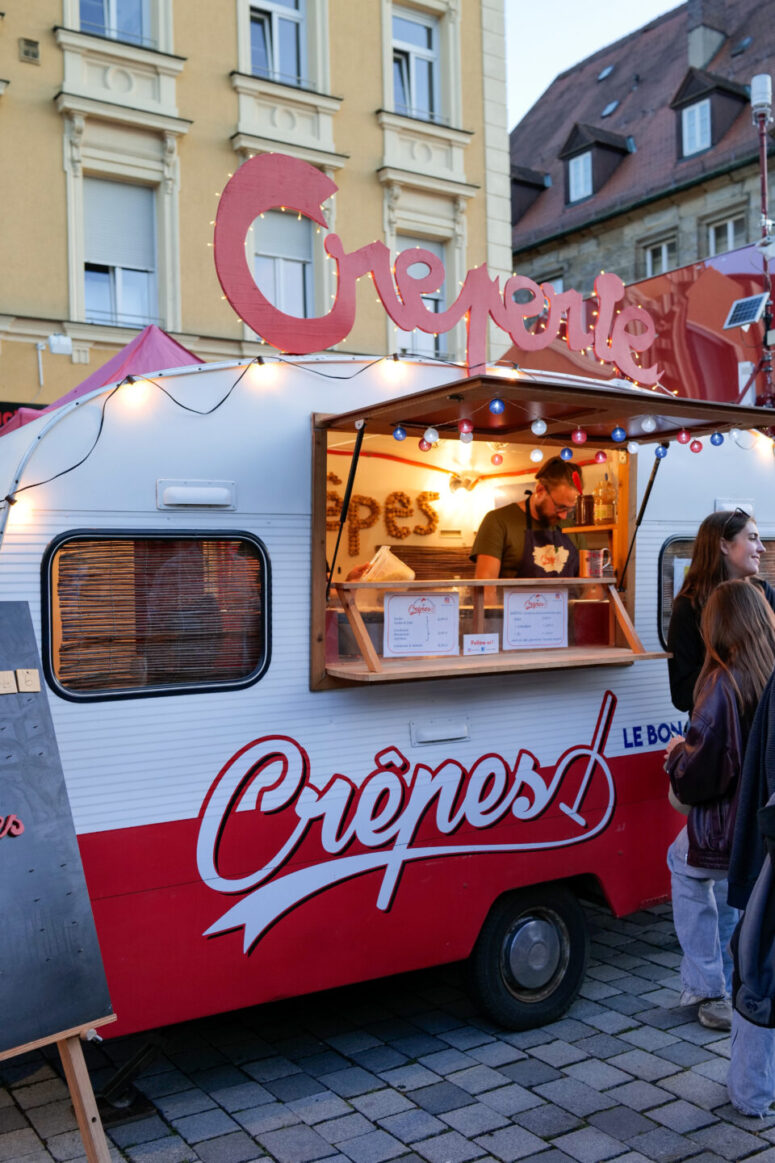 Ein Mann bereitet in einem rot-weißen Crêpes-Imbisswagen auf einem Markt unter freiem Himmel während des Bürgerfestes 2026 Speisen zu, während draußen Kunden warten. Der Wagen ist mit einem großen „Crêpes“-Schild versehen und steht vor bunten Gebäuden.