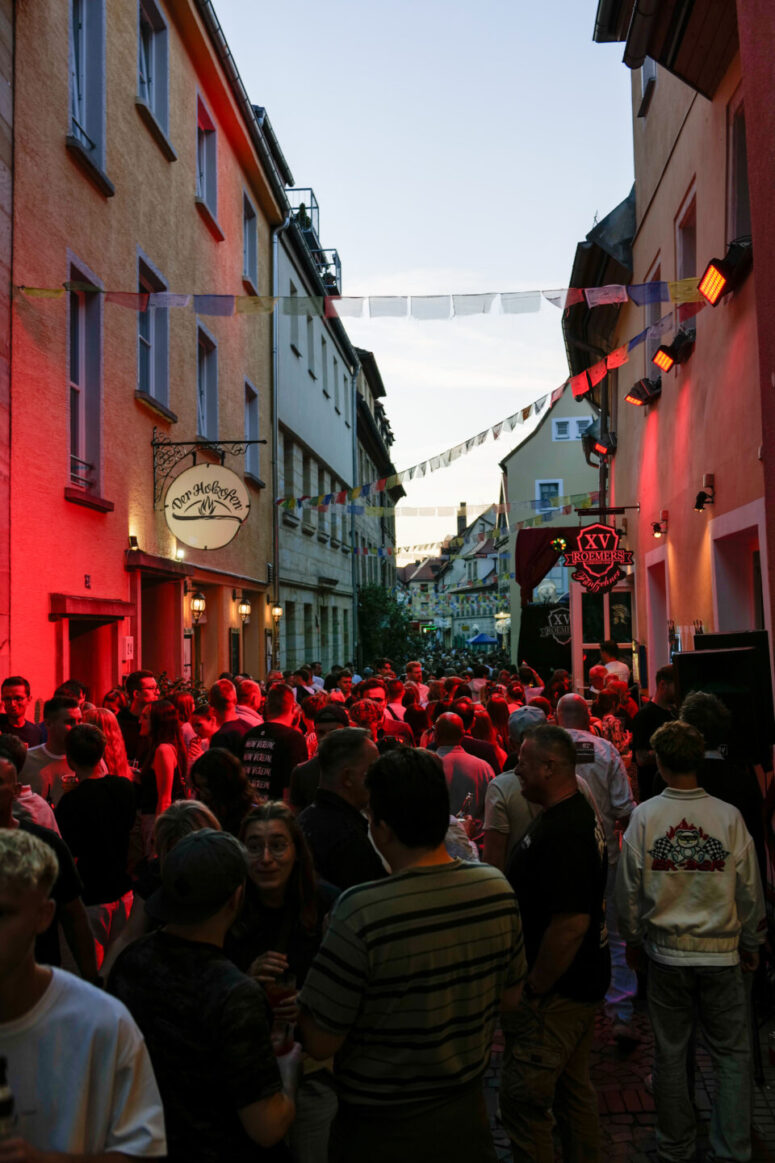 In einer schmalen Gasse, die mit hängenden Fahnen geschmückt und von bunten Lichtern beleuchtet ist, versammelt sich eine lebhafte Menschenmenge, während die Besucher in der Abenddämmerung zwischen den Gebäuden die festliche Atmosphäre des Bürgerfestes 2026 genießen.