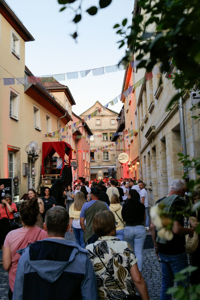Eine lebhafte Straßenszene zeigt eine Menschenmenge, die unter klarem Himmel durch eine schmale, mit Kopfsteinpflaster gepflasterte Gasse schlendert, die von bunten Fahnen, Terrassen und historischen Gebäuden gesäumt ist – und damit die festliche Stimmung des Bürgerfestes 2026 einfängt.