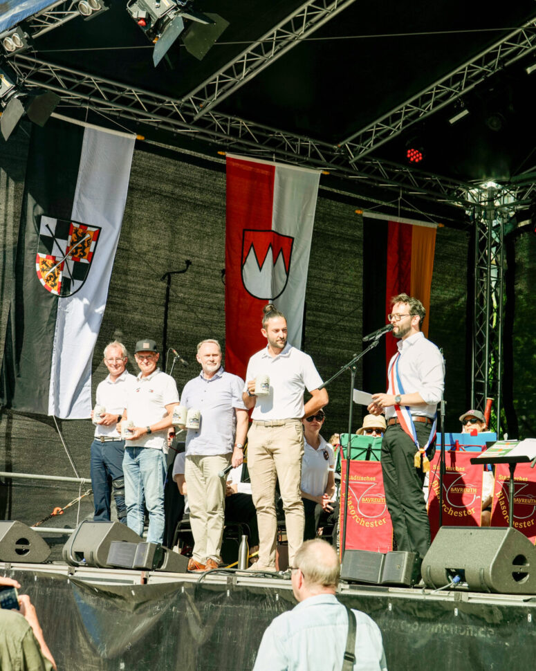 Auf dem Bürgerfest 2026 steht eine Gruppe von Männern auf einer mit regionalen Flaggen geschmückten Freiluftbühne. Ein Mann spricht ins Mikrofon, während andere Bierkrüge in den Händen halten; im Hintergrund spielt eine Band, und Zuschauer schauen zu.